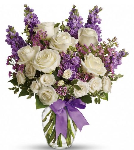 Lush white roses and purple stock mix with delicate lavender waxflower and green pitta negra for an elegant English garden look. The decorative bouquet is finished with a pretty satin ribbon. White roses meet lavender stock and waxflower in this lush, elegant arrangement. Presented in a classic clear glass vase with purple satin ribbon.