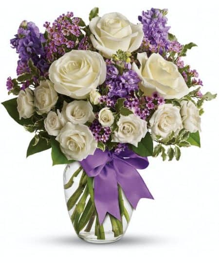 As enchanting as coming across a hidden cottage in the countryside! Lush white roses and purple stock mix with delicate lavender waxflower and green pitta negra for an elegant English garden look. The decorative bouquet is finished with a pretty satin ribbon. White roses meet lavender stock and waxflower in this lush, elegant arrangement. Presented in a classic clear glass vase with purple satin ribbon. Approximately 9" W x 16" H.