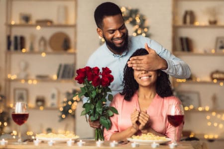 Smiling black man covering his woman eyes and giving her bunch of red roses, making surprise to beautiful lady. African american couple celebrating together at home or cafe